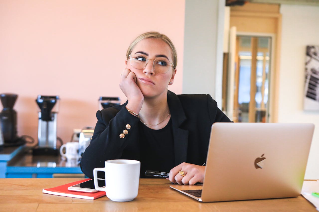 A young woman at a laptop in a cafe, pausing thoughtfully while researching online.