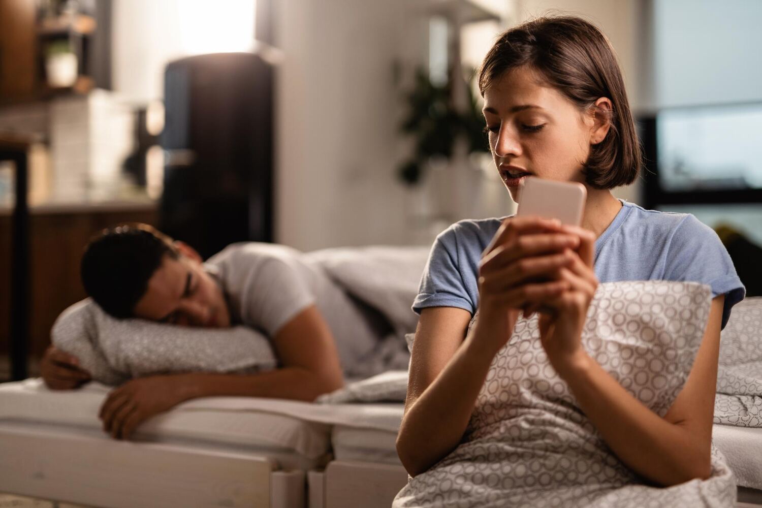 A woman sitting on the edge of a bed at night looking at her phone while her partner sleeps behind her.