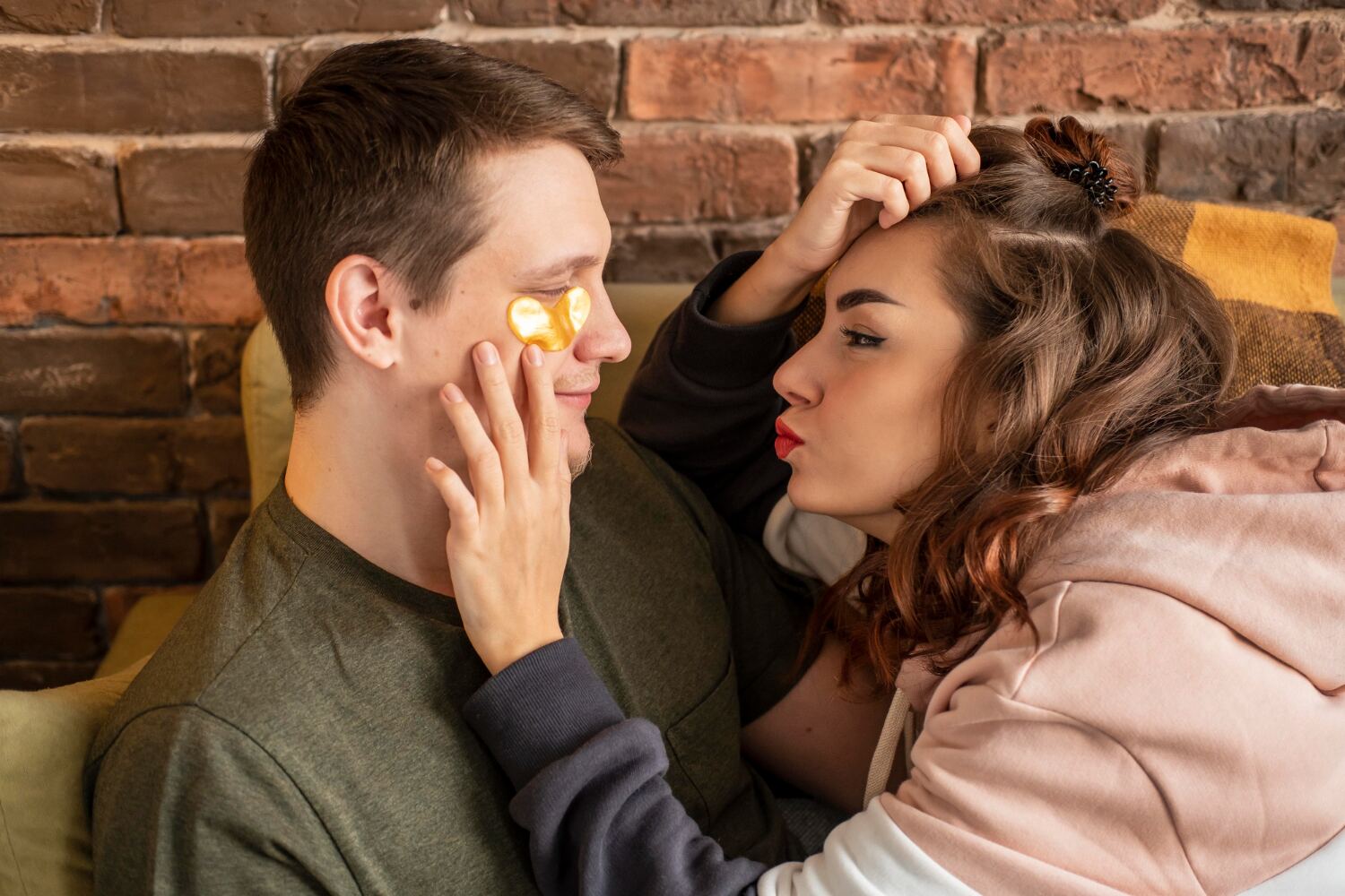 A young couple sitting close together on a couch in front of a brick wall, looking at each other quietly.