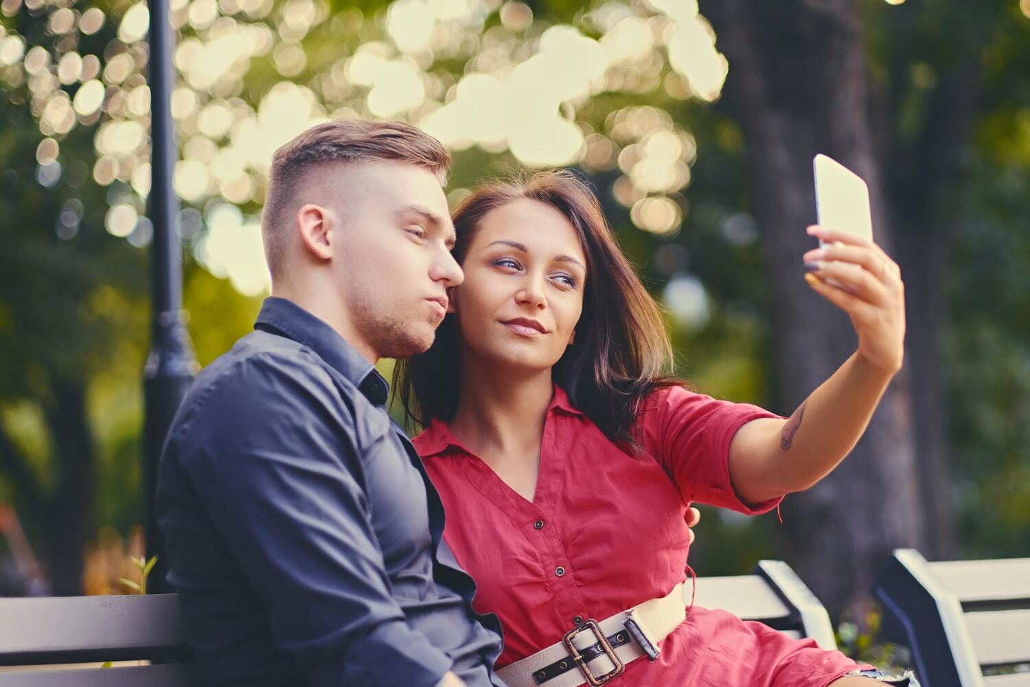 A couple sitting on a park bench taking a selfie together in warm afternoon light.