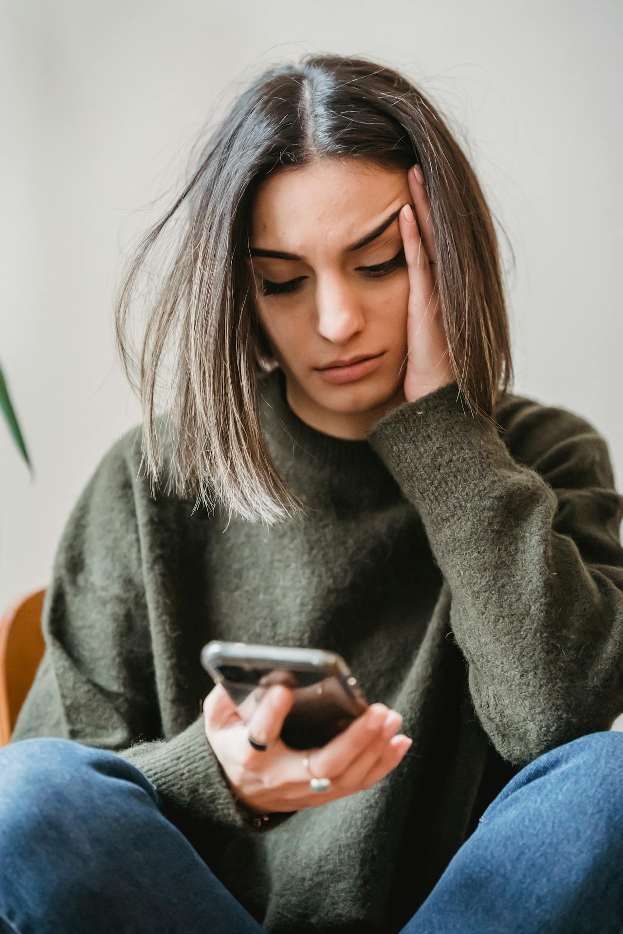 A young woman sitting on a chair, head in hand, looking at her phone with a worried expression.