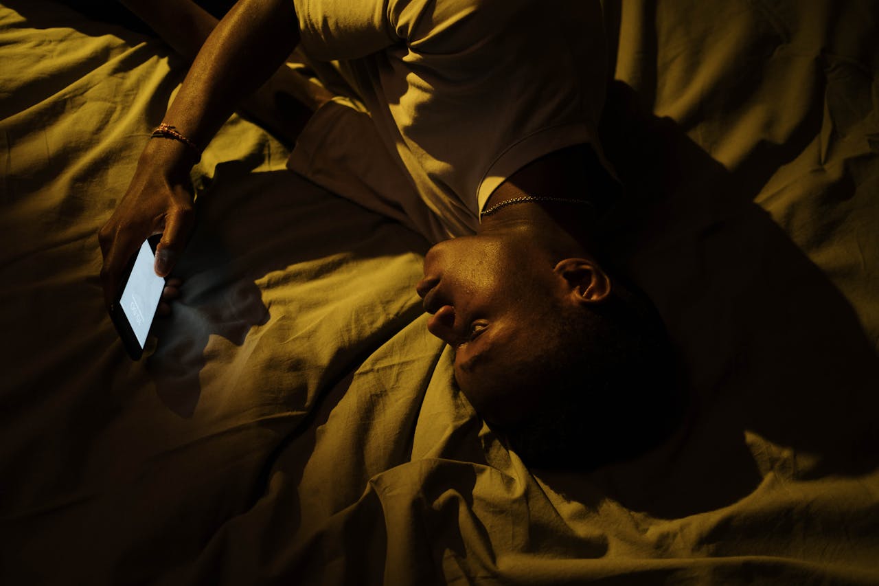 Overhead night shot of a person lying in bed, face dimly lit by the glow of a phone screen.