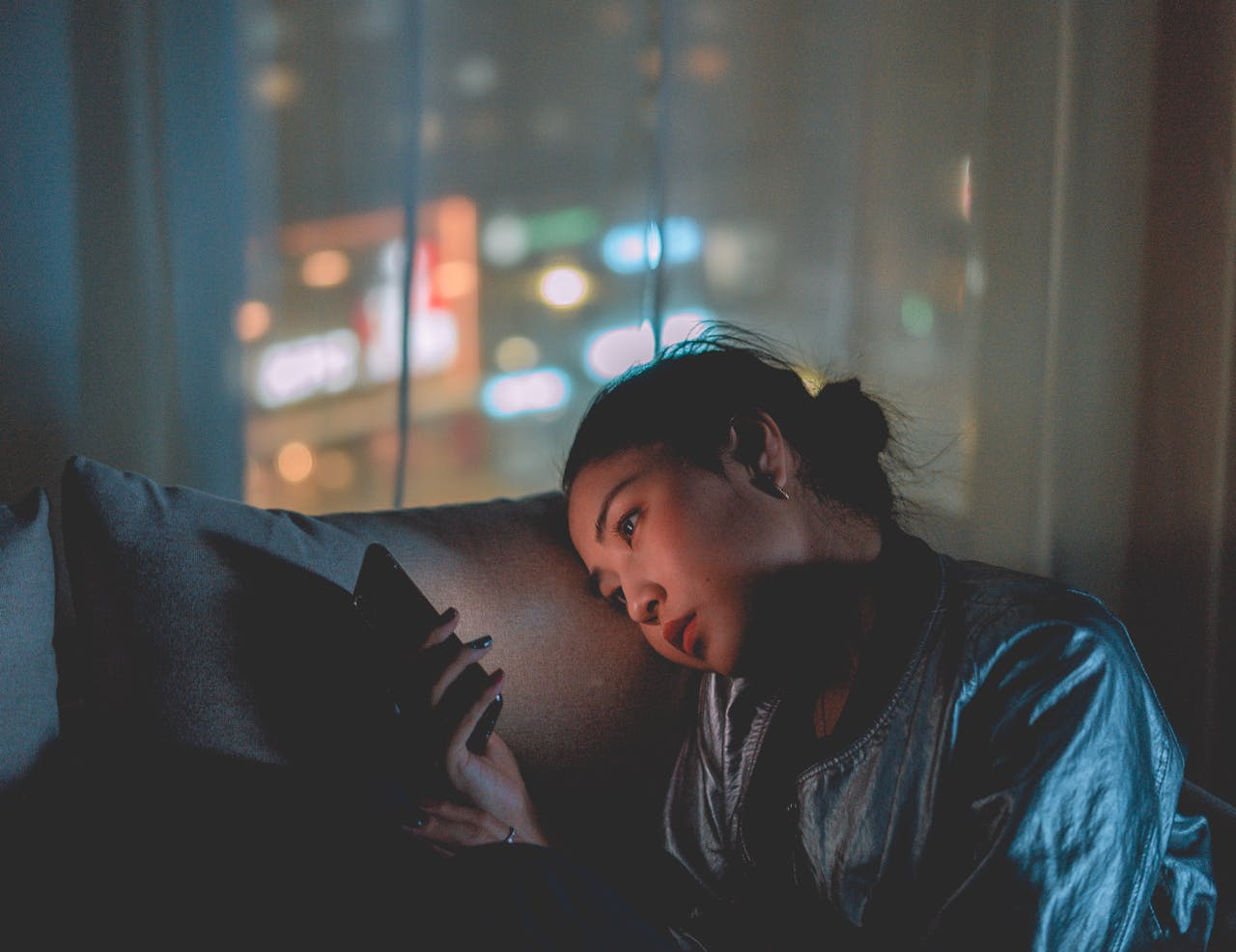 Young woman lying on a couch at night, face lit by the glow of her phone screen.