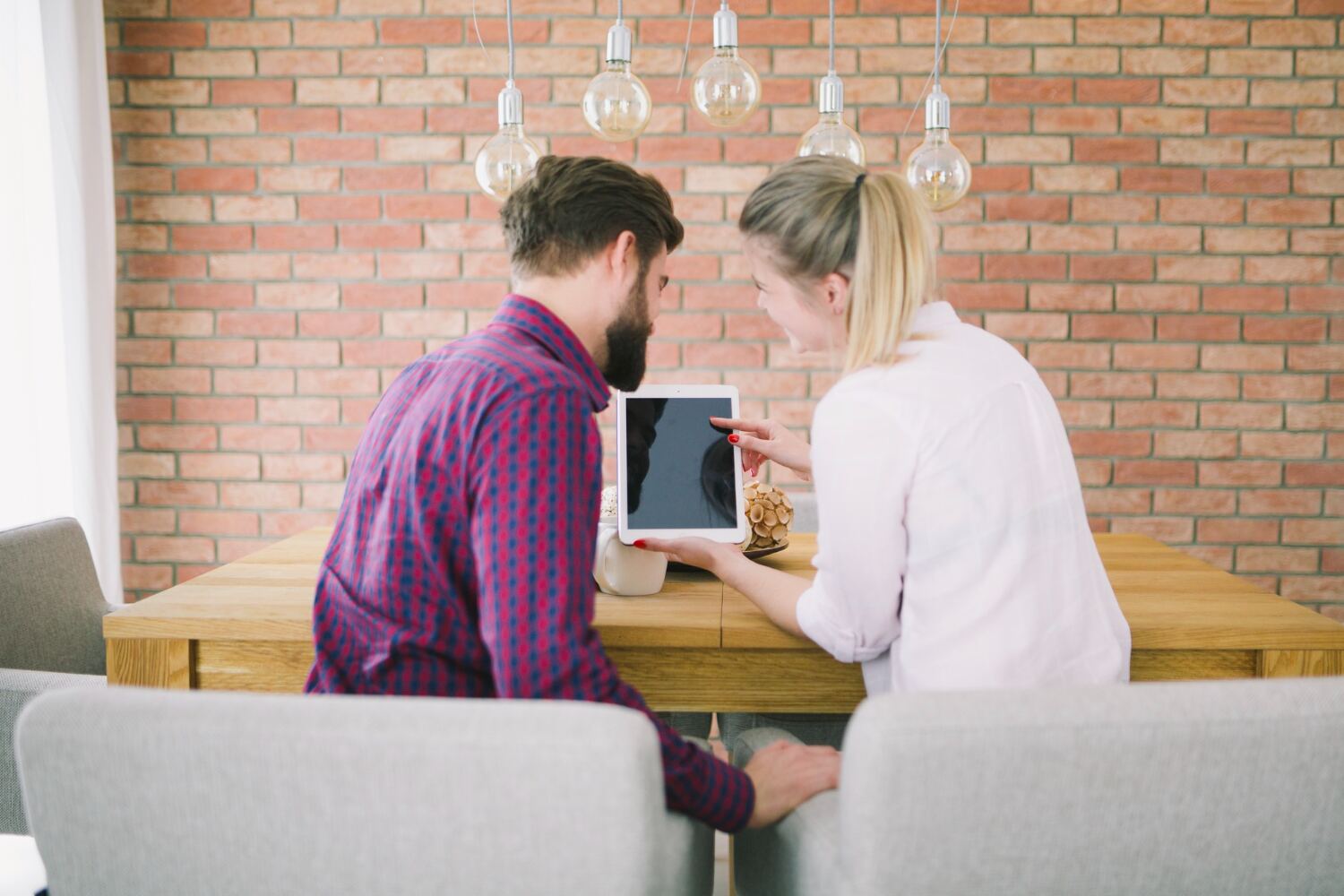 A couple sitting at a wooden table looking at a tablet together in front of a brick wall.