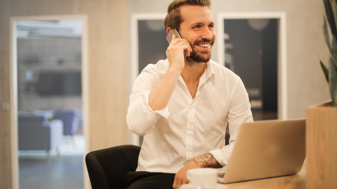 A person at a wooden desk holding a phone in one hand and typing on a laptop with the other, soft window light.
