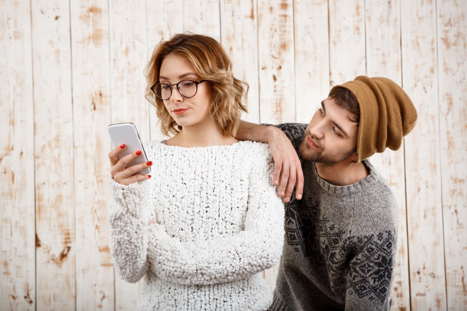 A woman in a white sweater looking at her phone while a man in a beanie leans on her shoulder watching the screen.